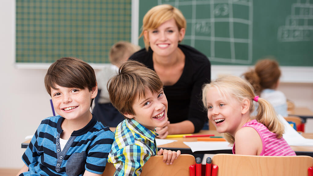 Zwei lachende Jungen und ein Mädchen im Grundschulalter sitzen im Klassenzimmer. Im Hintergrund die Lehrerin und eine Tafel.